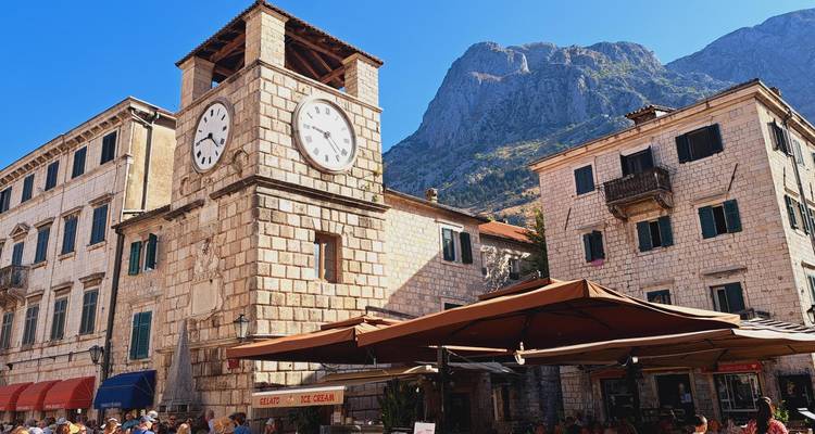 Torre de reloj de piedra y edificios históricos en una plaza antigua y soleada con empinadas montañas de fondo.