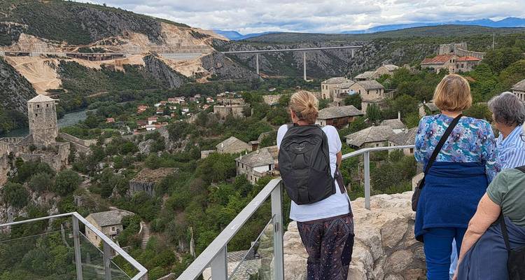 Viajeros mirando hacia un pueblo medieval en la ladera de una colina y un valle fluvial con un puente y montañas a lo lejos.