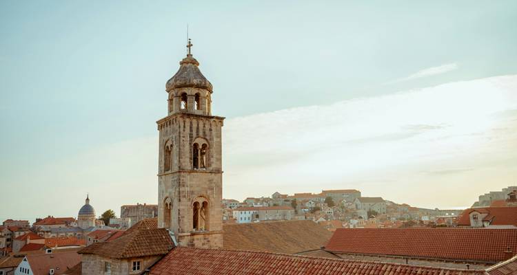 Torre de campanas elevándose sobre los tejados de terracota de una ciudad costera histórica bajo la cálida luz del atardecer.