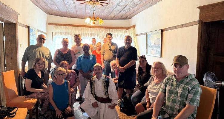Tour group poses indoors with a host in traditional Albanian costume inside a rustic village home