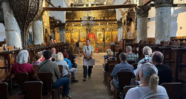 Local guide addresses visitors seated inside an ornate frescoed Orthodox church with gilded iconostasis