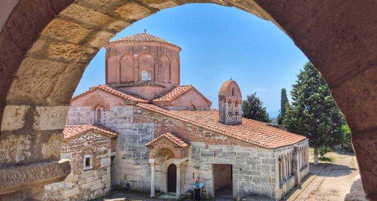 Historic Byzantine-style monastery framed perfectly by a stone arch, bathed in bright midday sun