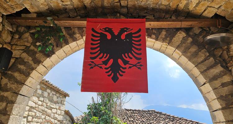 Albanian double-eagle flag hangs beneath a rustic stone archway with blue sky beyond
