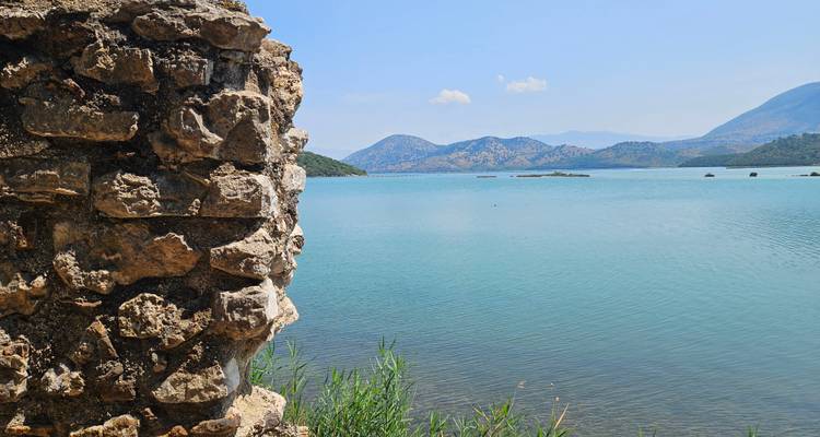 Tranquil blue lake with mountain backdrop viewed past the crumbling stone wall of an old fortress