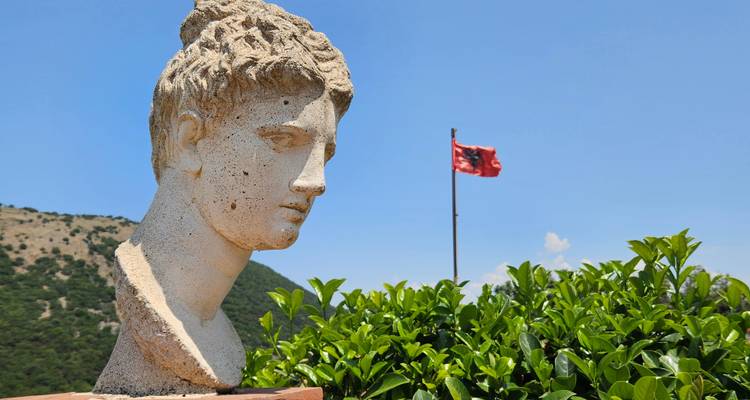 Stone bust of a classical figure with distant Albanian flag fluttering against a blue sky and green hills