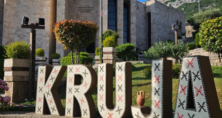 Dekorative große Textilbuchstaben, die "KRUJA" buchstabieren, in einem gepflegten Garten vor einem Steinmuseumsgebäude