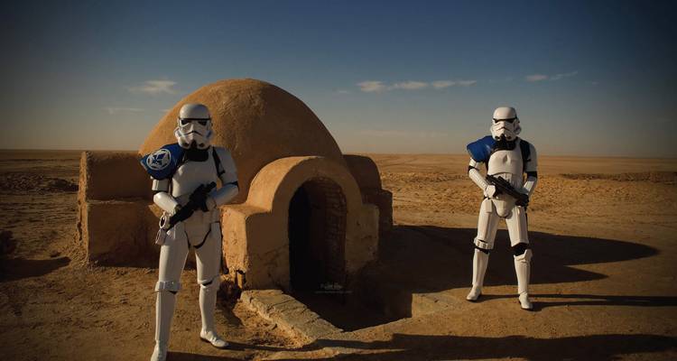 Personnages costumés debout dans un paysage désertique devant un bâtiment à coupole.