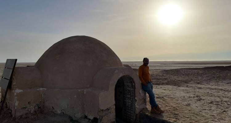 Une personne debout à côté d'une structure en dôme dans un désert au coucher du soleil.
