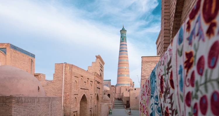 Vue plongeante sur une rue étroite vers le minaret rayé d'Islam Khodja encadré par des tissus colorés.