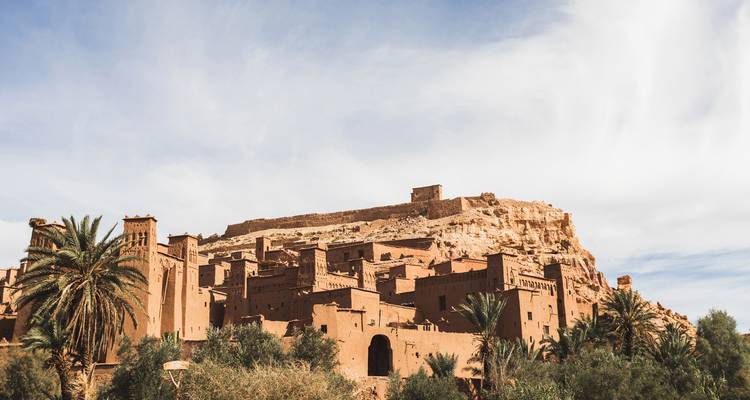 Iconic fortified village of Aït Benhaddou rising beside palms under dramatic sky.