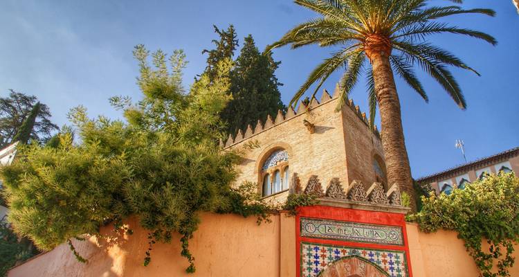 Building with intricate brickwork behind lush vegetation and palm trees.