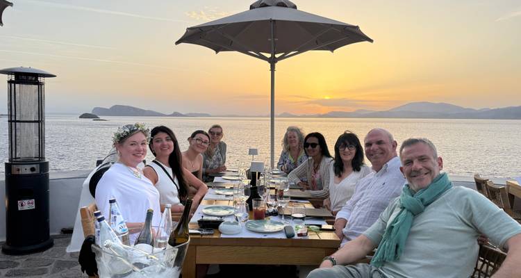 Repas de groupe à une table en plein air avec vue sur le coucher de soleil au-dessus de la mer.