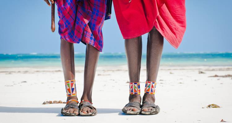 Onderste benen van twee Maasai-mannen in traditionele shukas die op een wit zandstrand aan de turquoise zee staan.