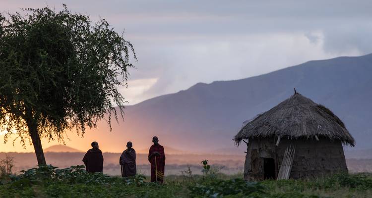 Drie Maasai-mannen in traditionele gewaden staan bij zonsondergang dicht bij een rieten hut met bergen op de achtergrond.