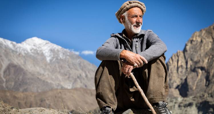 Elderly Hunza shepherd sitting on rock with walking stick against mountain backdrop