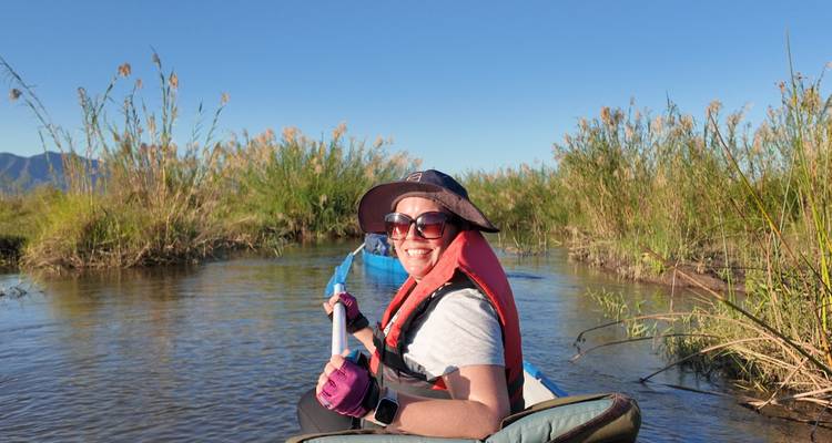 Een glimlachende kajakker met zonnebril en zwemvest peddelt door ondiep riet onder een blauwe lucht.