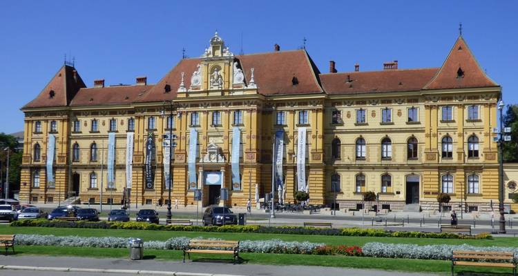 Neoklassiek gebouw op een stadsplein, blauwe lucht en bloemen.