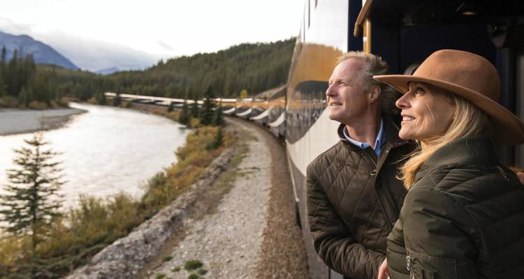 Smiling mature couple leaning from a luxury train to admire forested Rocky Mountain scenery along a river.
