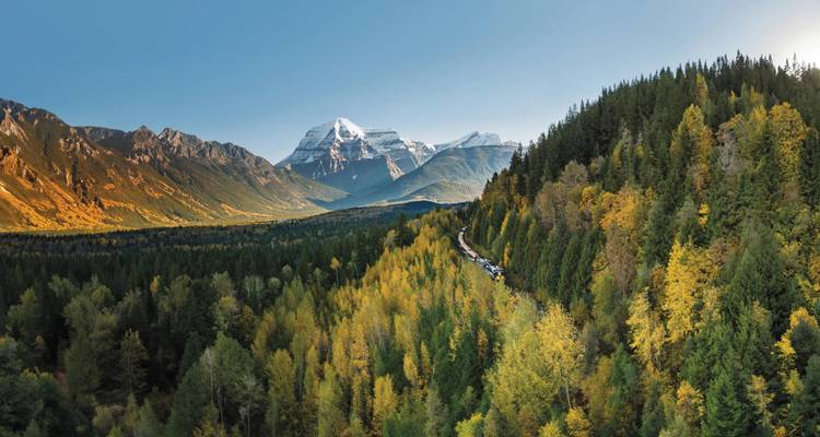 A passenger train snakes through a vast evergreen valley framed by snow-capped peaks in the Canadian Rockies.