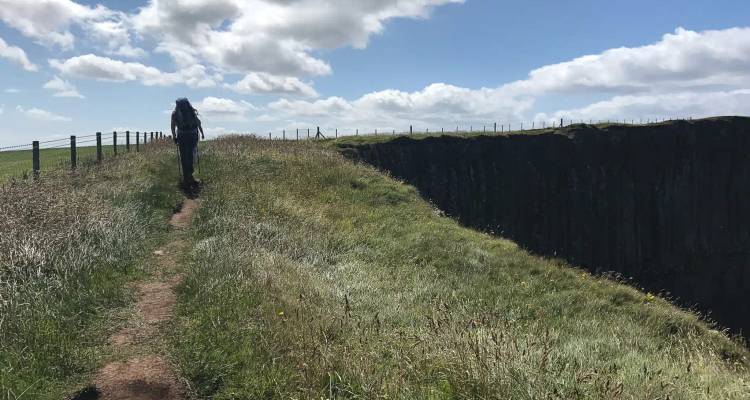 Backpacker loopt langs een graspaden wandelpad aan de rand van klif onder een lucht vol wolken.