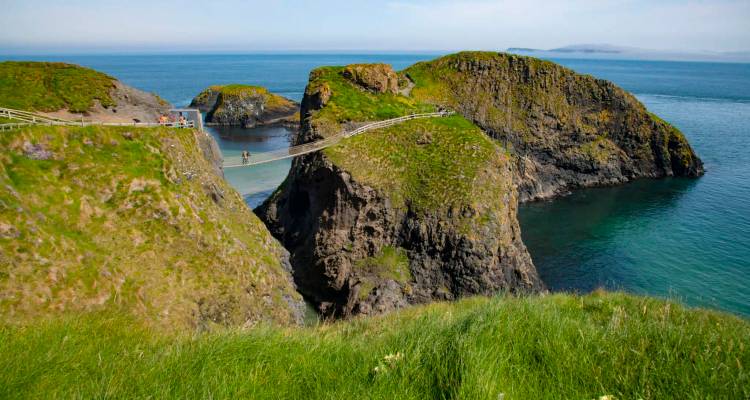 Iconic rope bridge linking grassy sea stacks above clear turquoise water with visitors crossing.