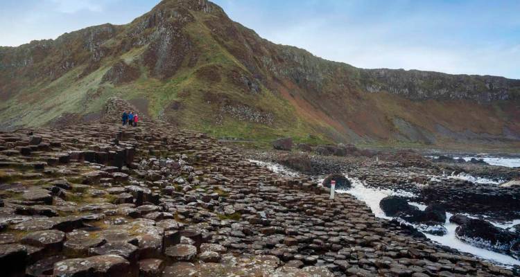 Visitors walking across the famous hexagonal basalt stones of Giant's Causeway beneath a rugged mountain.