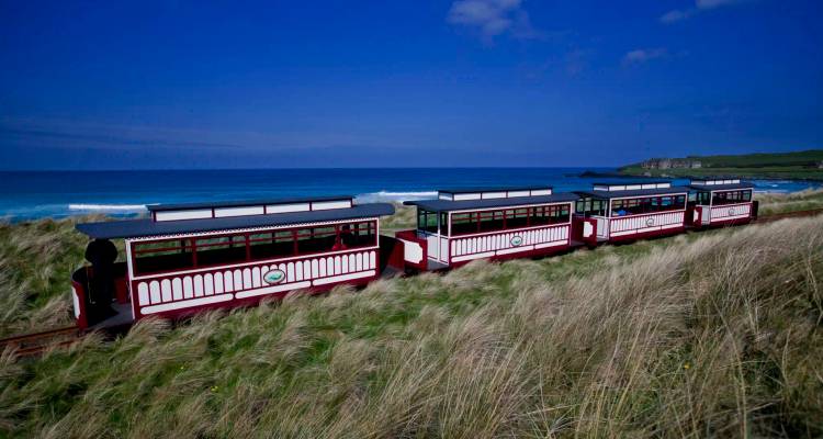 Red and white open-air tourist train travelling through tall coastal grasses beside a blue sea.