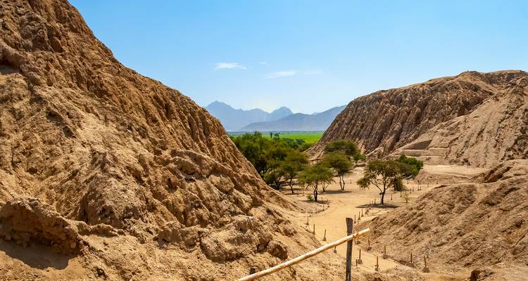Las colinas áridas de color marrón claro enmarcan un valle arenoso con acacias dispersas y campos verdes lejanos bajo un cielo azul despejado.