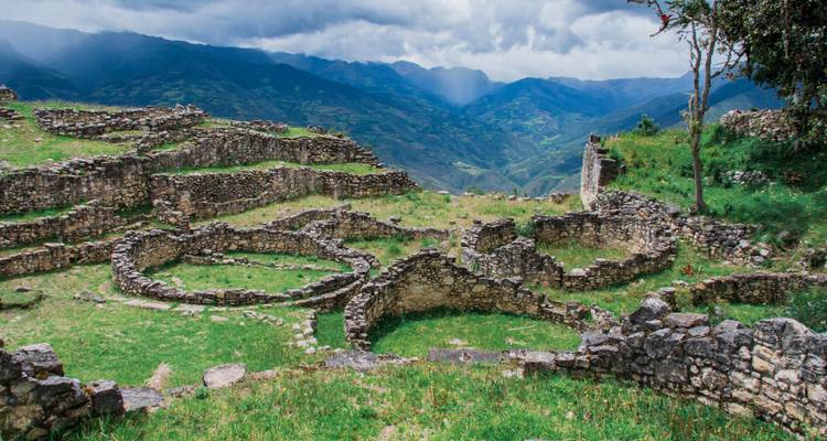 Muros de piedra y ruinas circulares de una antigua fortaleza en la cima de una colina que se alzan sobre verdes y exuberantes montañas bajo nubes espectaculares.