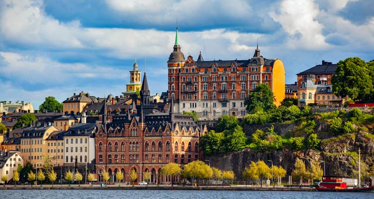 Historic brick and stone buildings with spires line Stockholm’s waterfront under a partly cloudy sky.