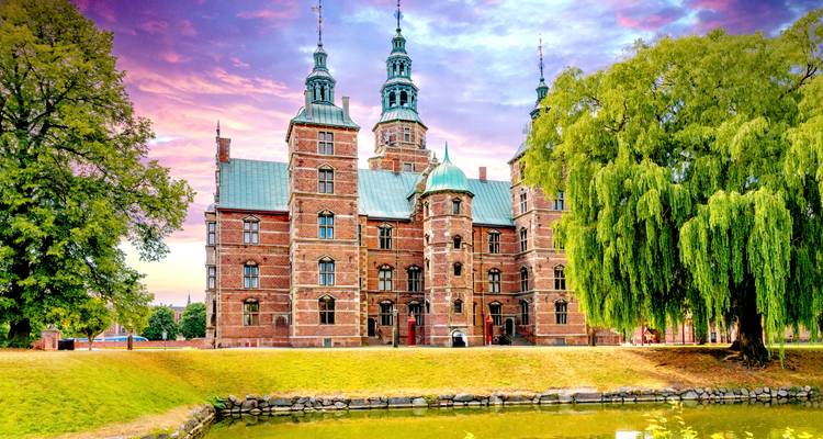 Rosenborg Castle, a red-brick Renaissance palace with turrets, stands beside manicured lawns and a reflective moat at sunset-tinted sky.