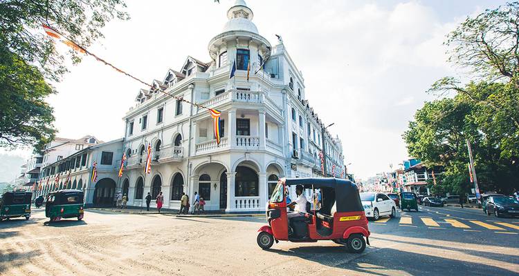 Tuk-tuk roulant devant un bâtiment de style colonial dans un environnement urbain ensoleillé.