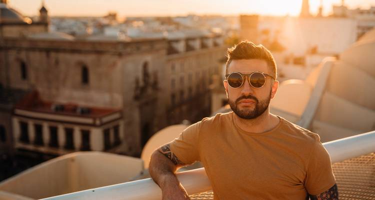 Homme élégant portant des lunettes de soleil posant au coucher du soleil au sommet du Metropol Parasol de Séville avec le paysage urbain flou derrière lui.