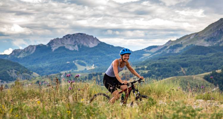 Eine lächelnde weibliche Radfahrerin fährt mit einem Mountainbike durch eine Wiese mit zerklüfteten Gipfeln und dramatischem bewölktem Himmel im Hintergrund.