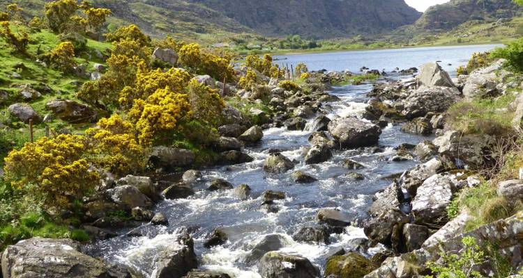 A fast-flowing stream cascades over boulders toward a tranquil lake set in a rugged valley.
