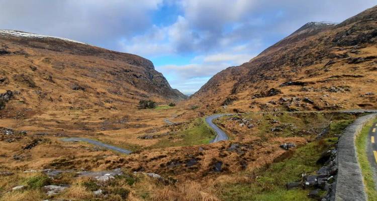 A winding road snakes through a dramatic brown mountain valley under a partly cloudy blue sky.