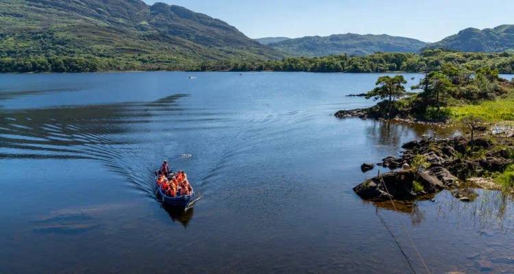 A small boat carrying tourists creates ripples across a tranquil lake surrounded by forested mountains.