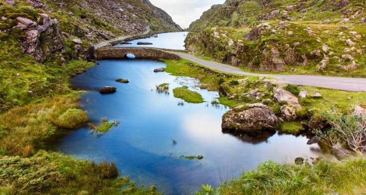 A stone arched bridge crosses a reflective stream amid rugged green mountain scenery.