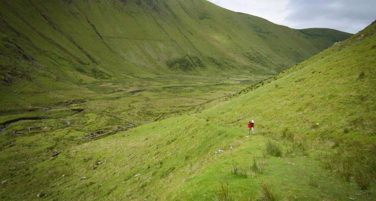 Single hiker in red makes their way along a vast green U-shaped valley.
