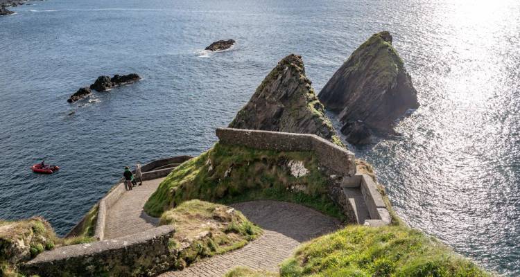Stone path spirals down to a small pier between two jagged rock stacks in deep blue sea.