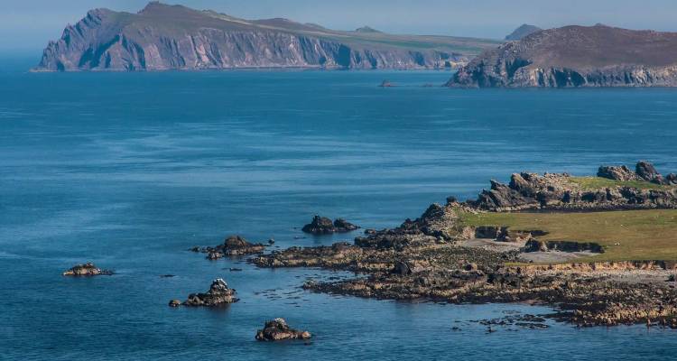 Distant headlands and rocky islets rise from a calm blue sea under hazy sunshine.