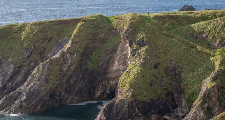 Steep green cliff with a lone figure standing at the crest, overlooking a secluded cove far below.