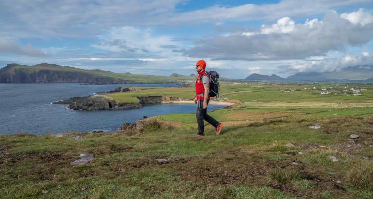 Backpacker in bright orange hat walks along a cliff-top path overlooking a sweeping bay and distant mountains.