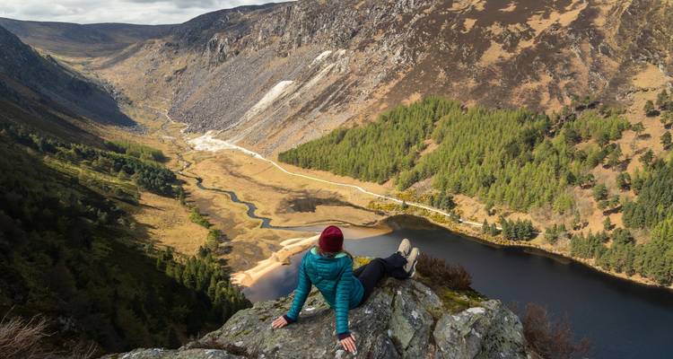 Person relaxing on a rocky cliff overlooking a valley and lake.