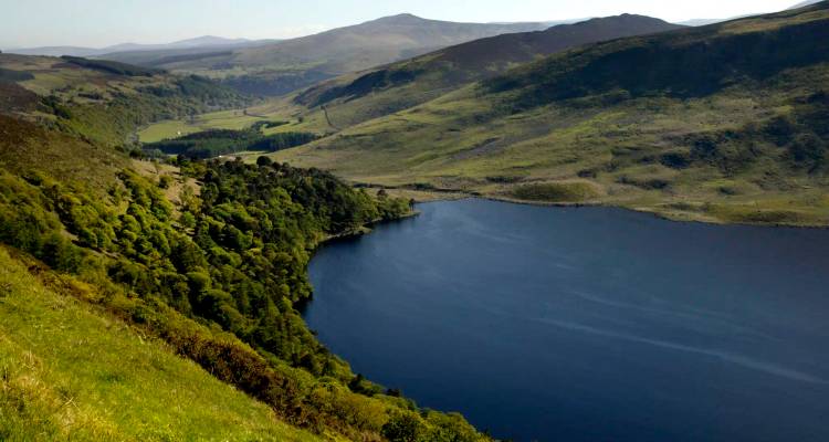 Deep blue lough bordered by lush green slopes and forested ridges, viewed from a high mountain viewpoint.