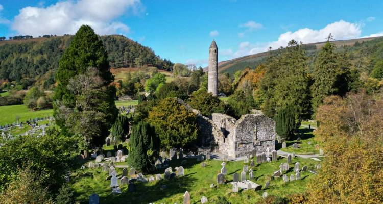 Historic monastic round tower rising above ancient stone ruins and a green cemetery set in a wooded valley.