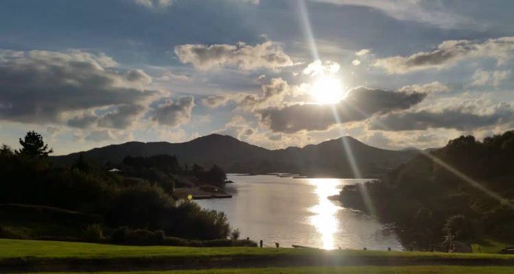 Low sun shining through dramatic clouds over a reflective lake framed by hills.