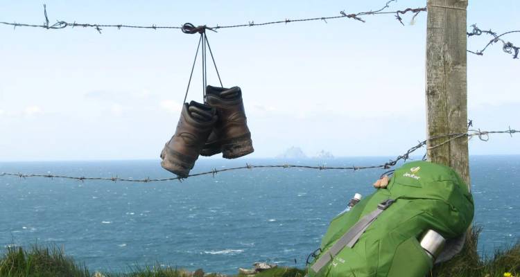 A pair of hiking boots hang on barbed wire above a blue sea with distant rocky islands, backpack leaning on post.