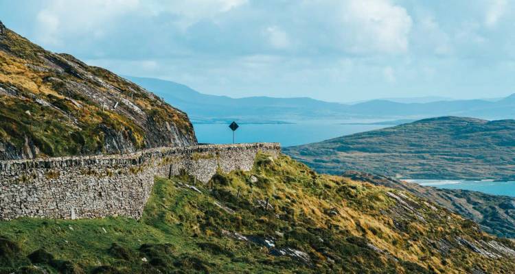 Stone wall road curving along a steep green cliff with wide ocean views and distant mountains.