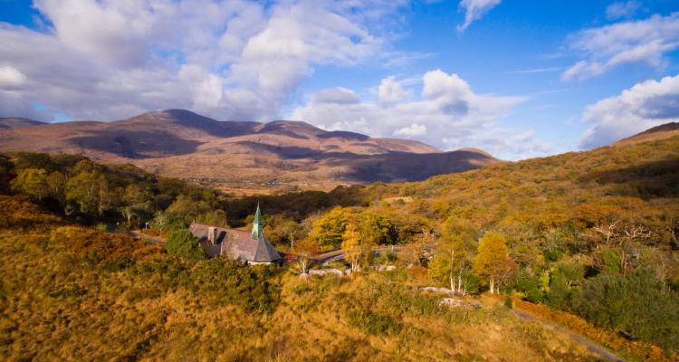 Autumn-coloured valley with a small church and spire, set beneath rolling mountains and a vibrant blue sky.